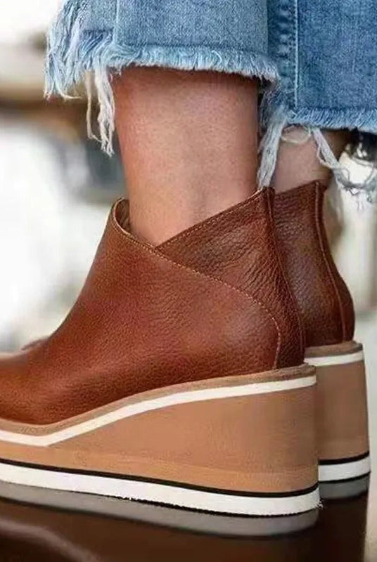 Lifestyle image of a woman sitting on a bench, wearing brown Elara casual wedge ankle boots, looking relaxed.