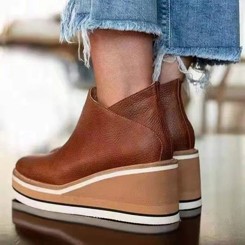 Lifestyle image of a woman sitting on a bench, wearing brown Elara casual wedge ankle boots, looking relaxed.