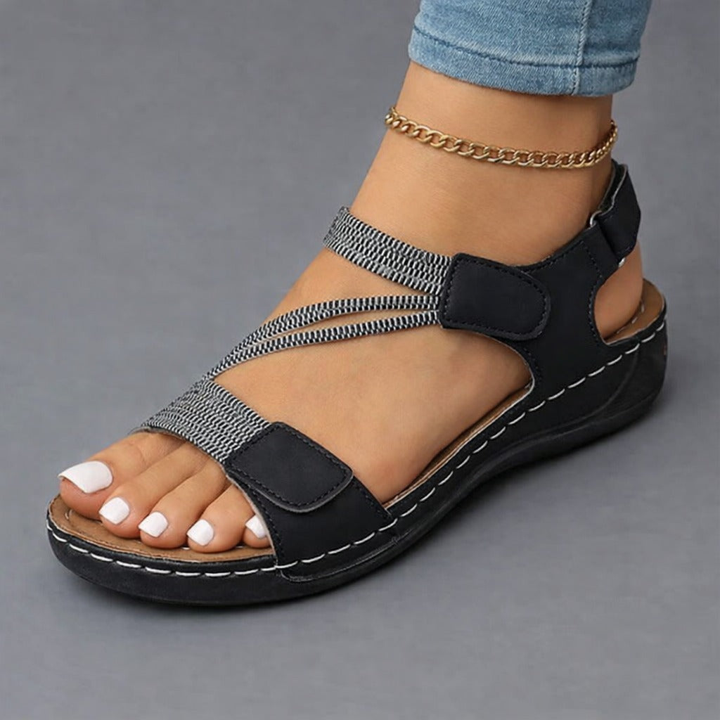 A woman's feet wearing beige Liana orthopedic comfort sandals, standing on a cobblestone path, demonstrating natural walking motion and flexible fit.