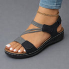 A woman's feet wearing beige Liana orthopedic comfort sandals, standing on a cobblestone path, demonstrating natural walking motion and flexible fit.